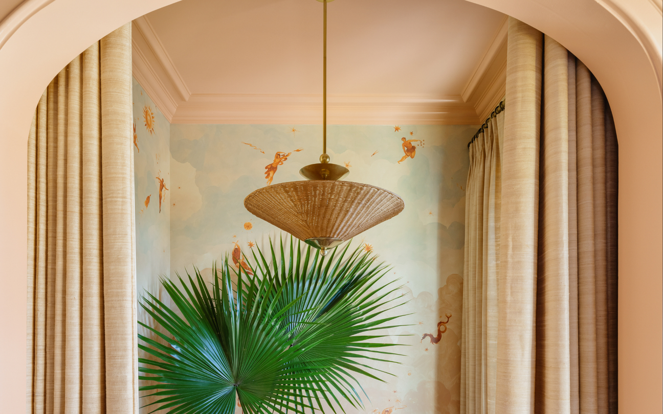 Woven pendant light over a fan palm in an arched peach alcove with a blue celestial mural and beige curtains