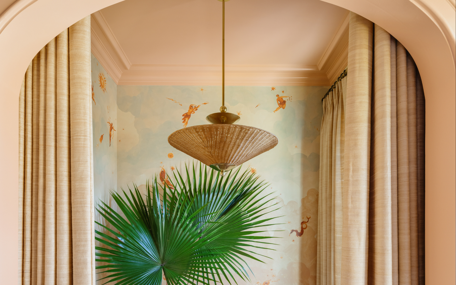 Woven pendant light over a fan palm in an arched peach alcove with a blue celestial mural and beige curtains