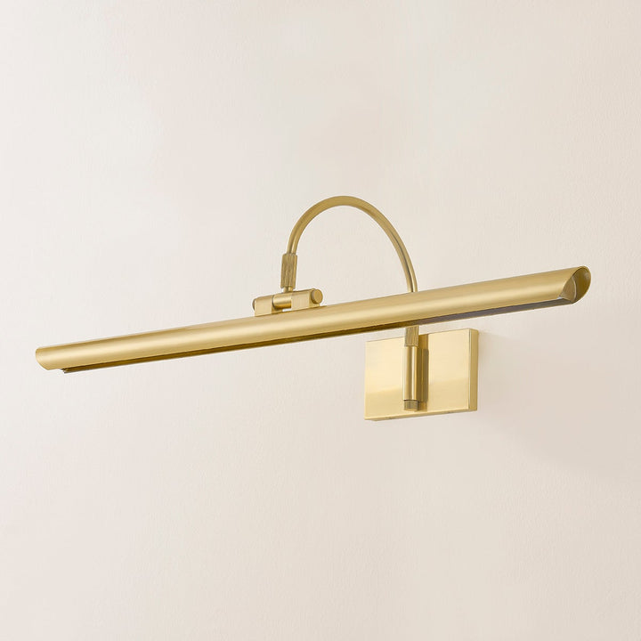 Front view of Elowen Picture Light showing slim antique gold bar lamp with curved arm and square wall mount on pale wall