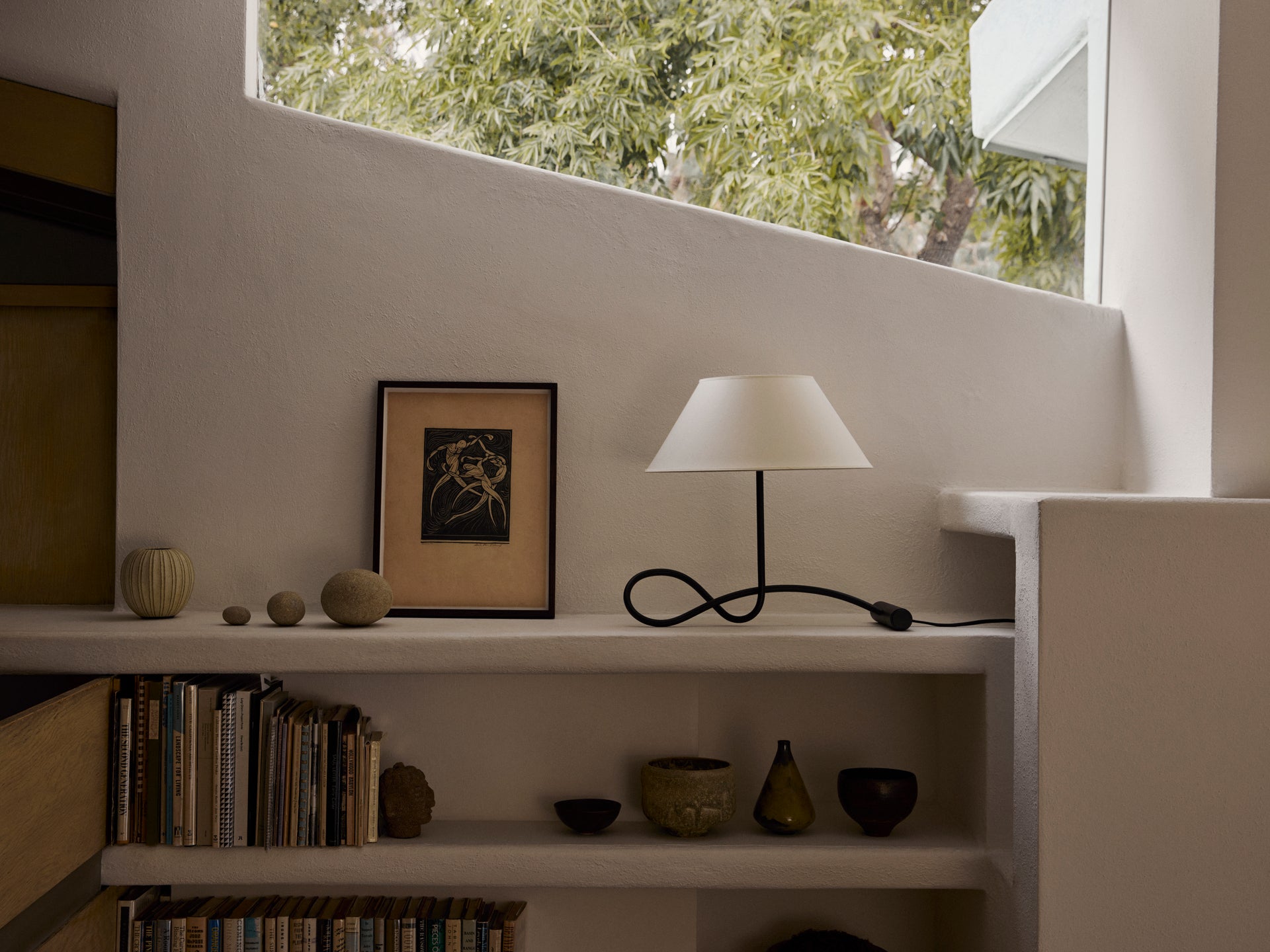 Calabria table lamp on low stucco shelf beside framed print and stone spheres beneath a slanted window with trees outside
